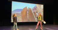 Shaelyn Silverman and Juliet Ryan-Davis MC ReelRock 14, in front of a slideshow showing people bouldering in a crack in boulder in Bishop CA.
