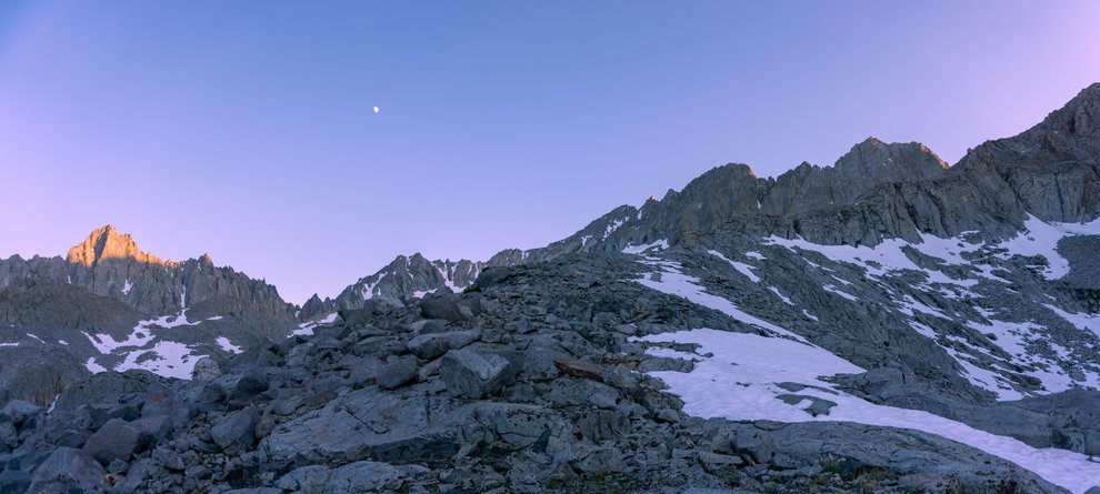 Photo of the high Sierra crest peaks with the moon above, and a little bit of remaining alpenglow on the highest peak and a patchy snow apron at dusk.