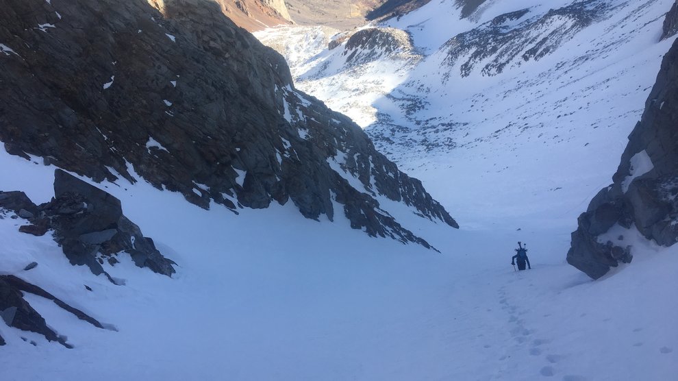View of a narrow, snow-filled couloir and a skier hiking it up.