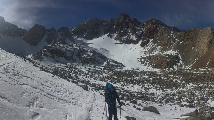View of rocky but snow-covered mountain terrain. A skier is in the foreground. The sun is shining.