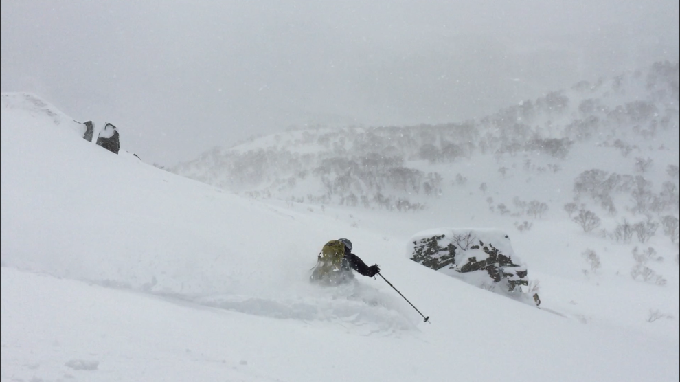 View of a skier rieding down a slope in deep snow, a hillside is visible in the background.