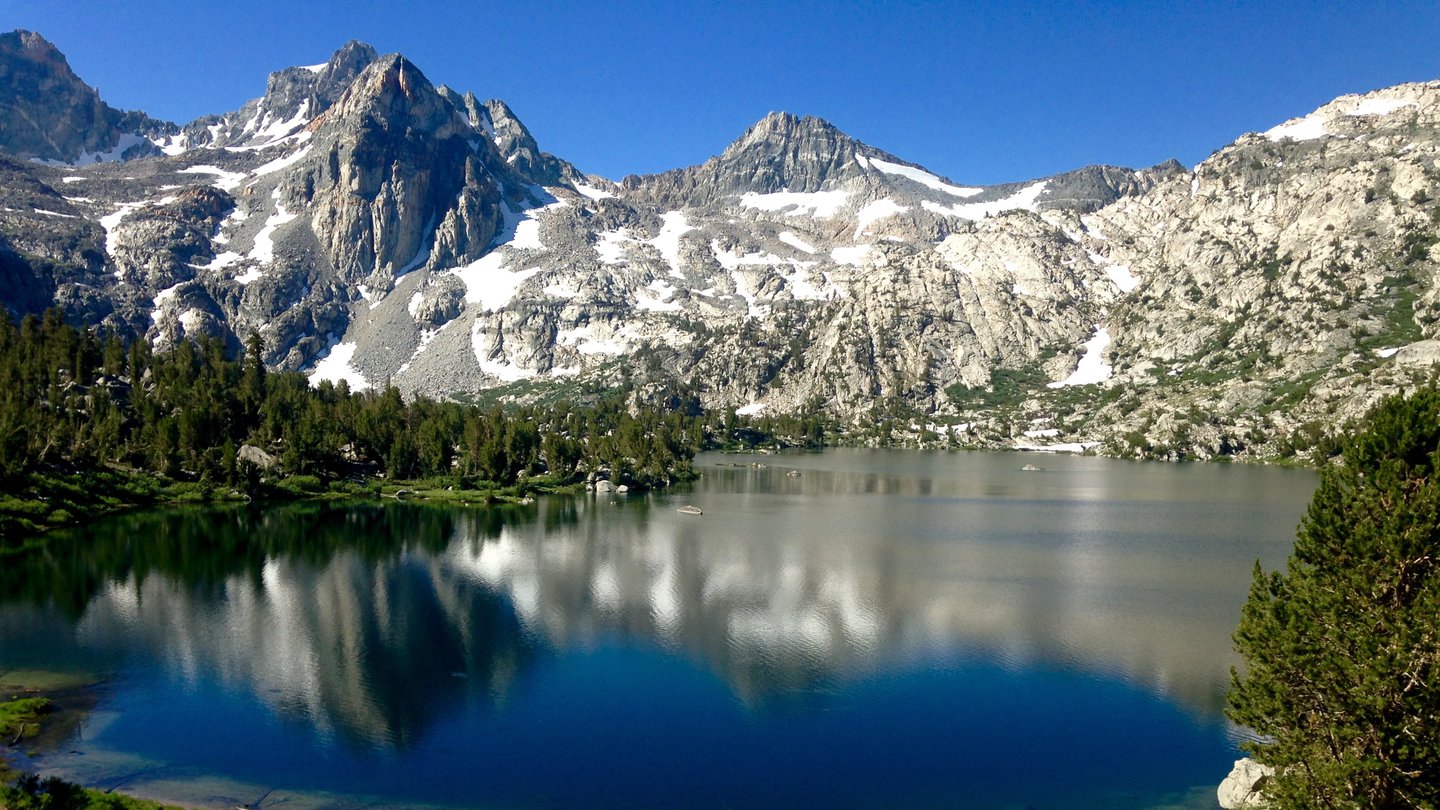 Blue, reflective Rae Lakes in the foreground surrounded by pine trees, stark gray striped peaks of the Sierras in the background.