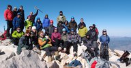 A large group of people from the Caltech Alpine Club posing among rocks at the summit of San Gorgonio mountain. A clear blue sky stretches out in the background, and the group stands on a slight slant, sloping left to right.