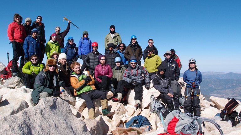 A large group of people from the Caltech Alpine Club posing among rocks at the summit of San Gorgonio mountain. A clear blue sky stretches out in the background, and the group stands on a slight slant, sloping left to right.