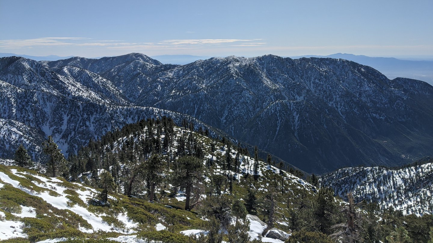 View down the valley from the hike on Mt Baldy. The ground is still half-covered in snow, and scarce treeds cover the hillsides. Blue skies.