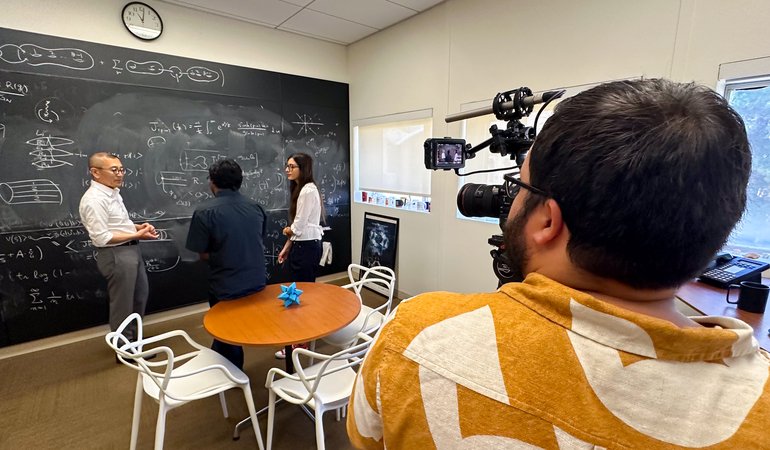 three people stand at blackboard with foreground person holding camera filming