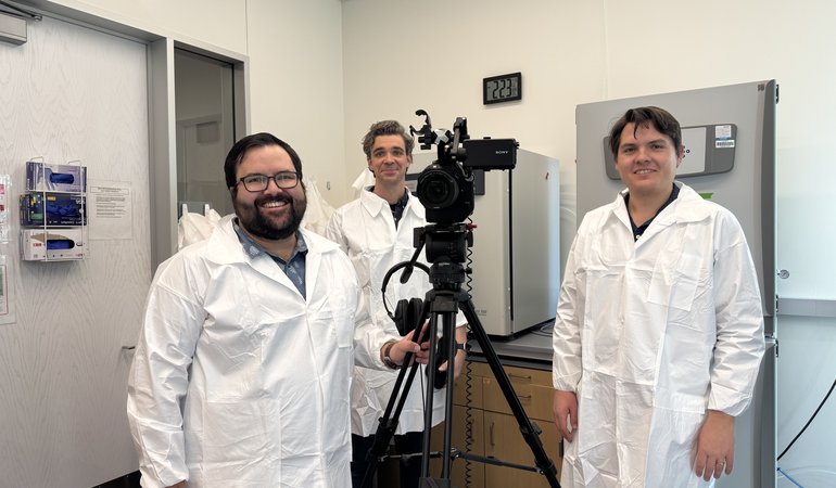 Three men in lab coats stand next to a camera.