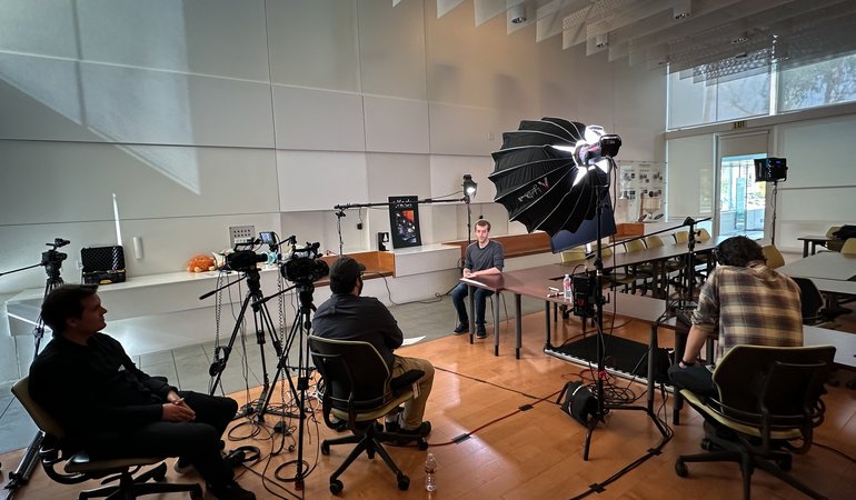 A man sits in the center of a large room for an interview, surrounded by lights, a director, a sound mixer, and a camera operator.