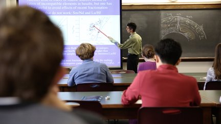 teacher pointing with stick to projected image students in foreground