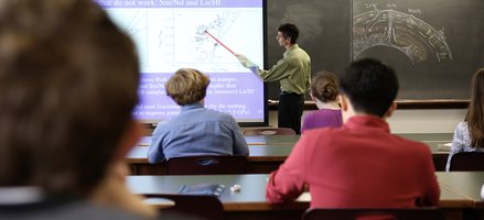 teacher pointing with stick to projected image students in foreground