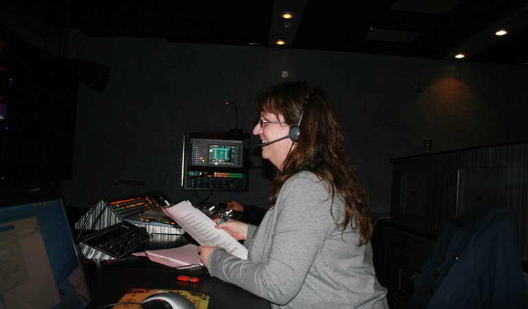 woman wearing headphones smiles and directs a broadcast from behind a video switcher
