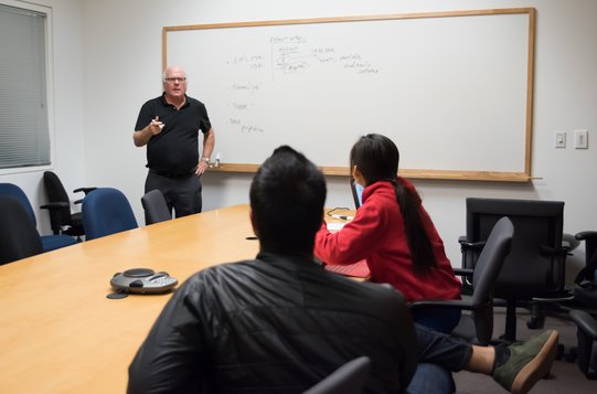 Camerer standing at a white board talking to two students
