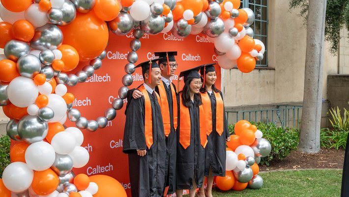 A group of four students in graduation regalia standing in front of a Caltech-themed backdrop decorated with orange, white, and silver balloons