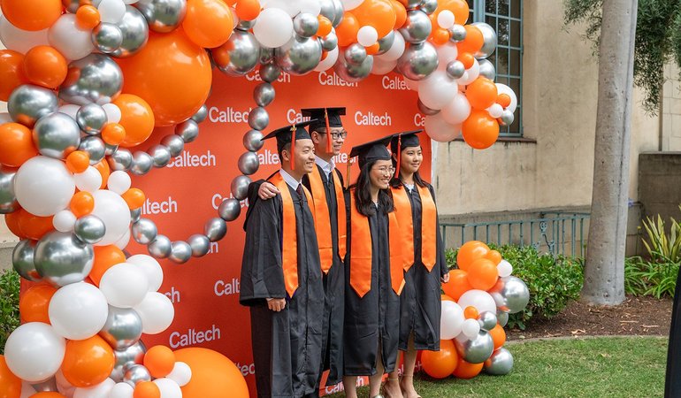 A group of four students in graduation regalia standing in front of a Caltech-themed backdrop decorated with orange, white, and silver balloons
