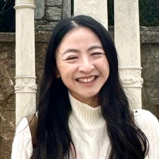 Woman with long dark hair wearing white turtleneck sweater. She is outside and there are three white columns behind her.