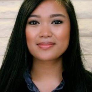 Woman with long dark hair against a white wall. She is wearing a dark blue shirt and black jacket