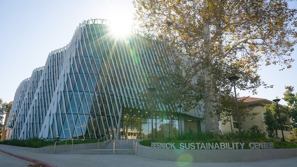 A building with a curved glass facade and diagonal shade fins on the exterior is shown, illuminated by the sun, with a sign reading Resnick Sustainability Center in silver letters in the foreground