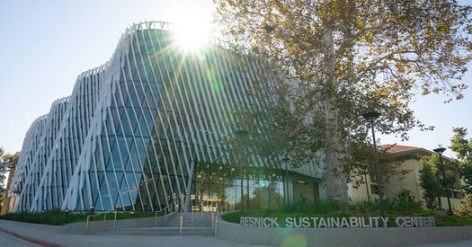 A building with a curved glass facade and diagonal shade fins on the exterior is shown, illuminated by the sun, with a sign reading Resnick Sustainability Center in silver letters in the foreground