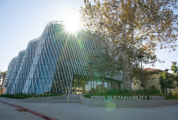 A building with a curved glass facade and diagonal shade fins on the exterior is shown, illuminated by the sun, with a sign reading Resnick Sustainability Center in silver letters in the foreground