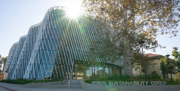 A building with a curved glass facade and diagonal shade fins on the exterior is shown, illuminated by the sun, with a sign reading Resnick Sustainability Center in silver letters in the foreground