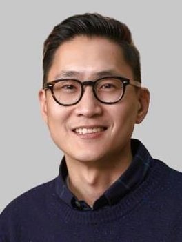 Man with dark hair and dark-rimmed glasses wearing a dark navy collared shirt and sweater