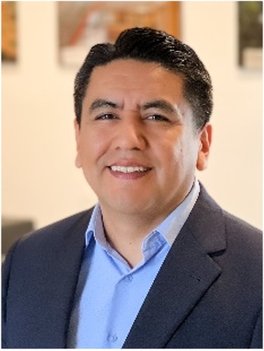 Man with dark hair wearing blue shirt and dark jacket. There is a pale orange building behind him.