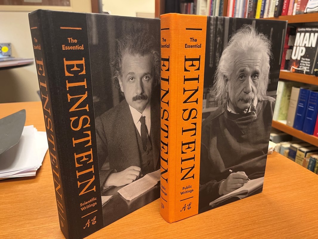 2 books, uprirght on a desk, bookshelf in background. Books are The Essential Einstein. Scientific Writings cover is of youthful Einstein. The Public Writings cover is a photo of an elderly  Einstein.