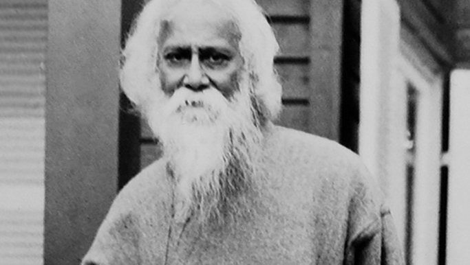 Greyscale image. Bengali poet Rabindranath Tagore stands outdoors, wearing a long sleeved robe like garment.