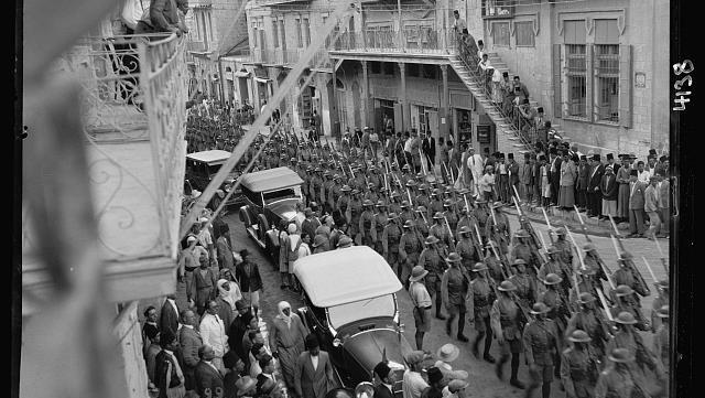 Black and white photo taken from a first floor balcony or window. Image is of British troops marching down street in Jerusalem with bayonets, alongside cars. Local people line the street observing the scene.