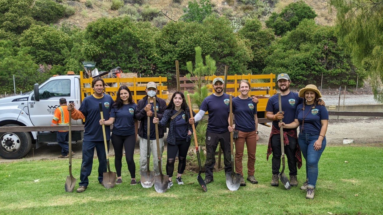 A group of people in dark blue shirts and dark long pants are standing on a grassy area. A hill and white truck are behind them.  They are holding shovels.