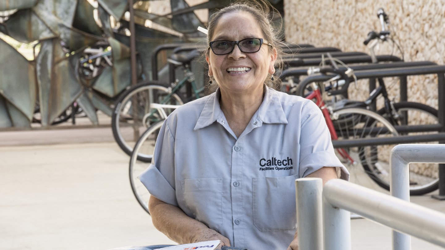 woman in light tan short-sleeved shirt. She is smiling and wearing glasses with black rims. She has a large garbage receptacle in front of her filled with used paper towels