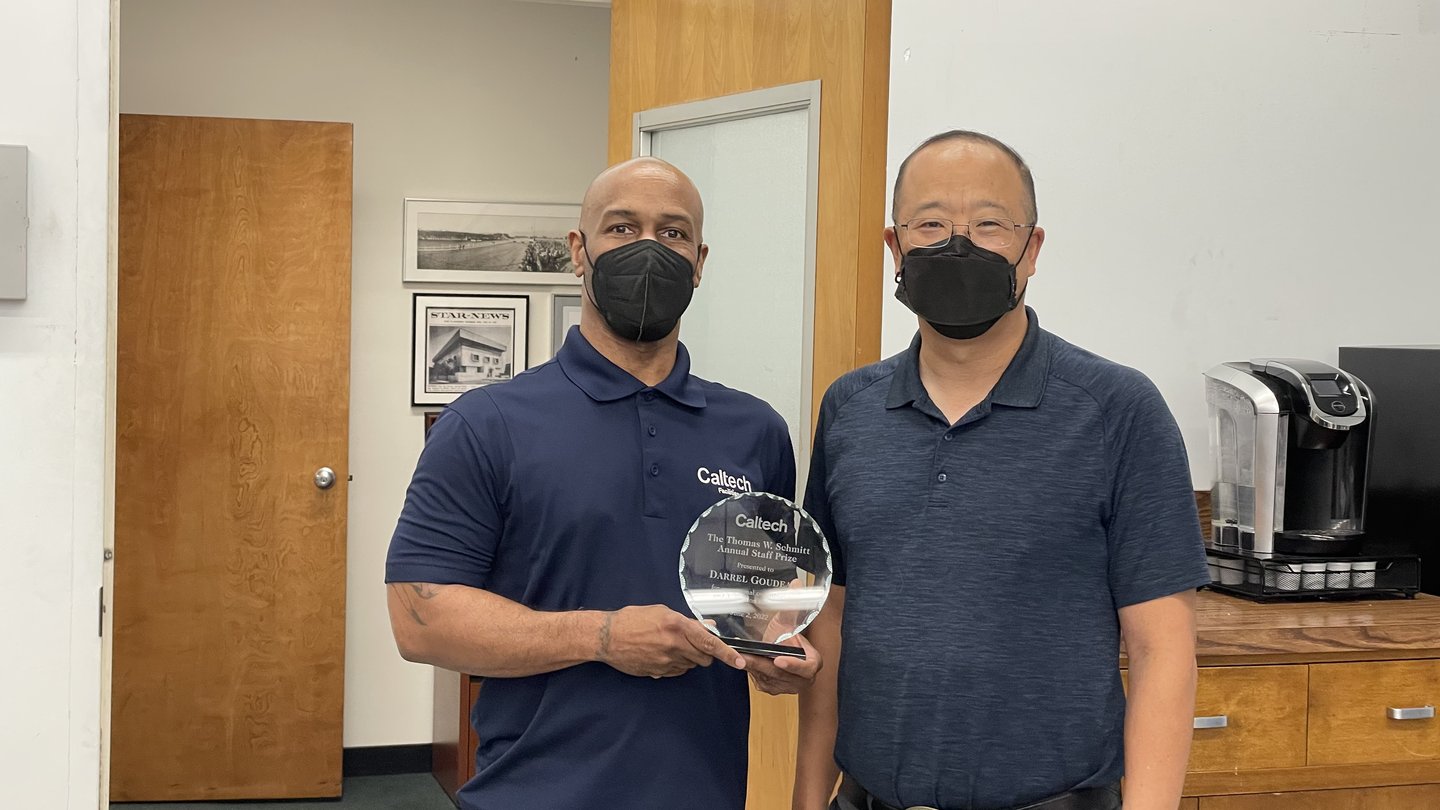 two men in dark blue shirts with collars. Both men are wearing black masks. One man is holding a round award with engraving