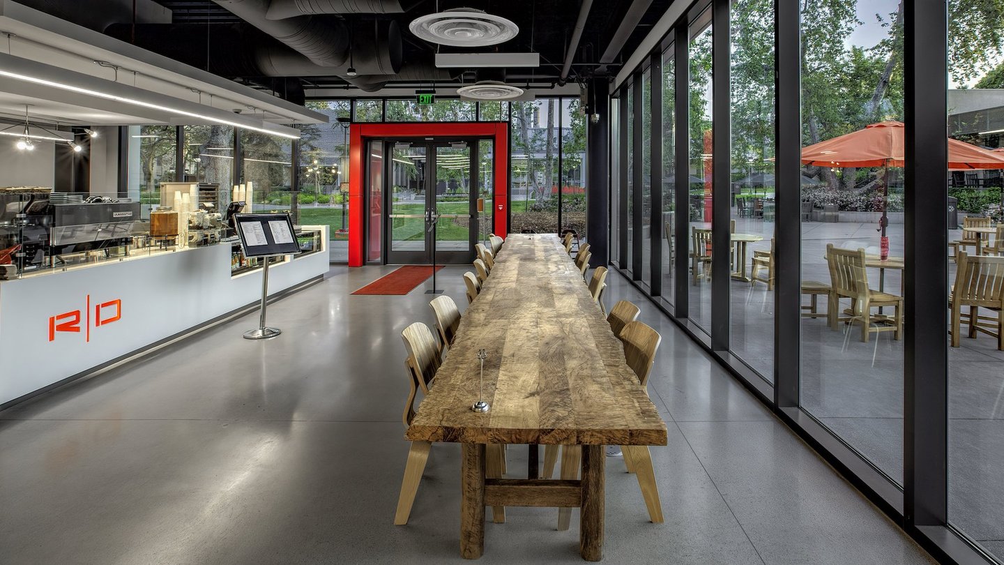 Long wood table in Hameetman Center made from Engelmann Oak. There are 6 chairs on each side of table. At the back is a red door frame denoting the "red door cafe" and on the left is the kitchen where workers prepare light lunch fare.