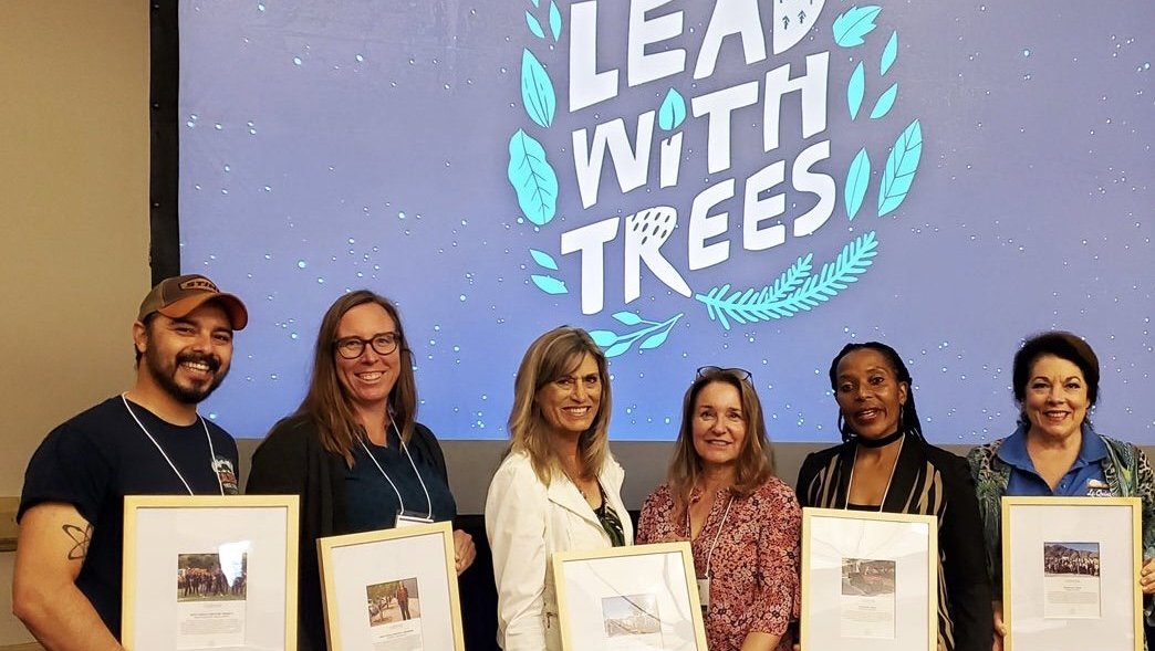 Six individuals are holding framed awards. There is a blue presentation slide projected on the wall behind them that says "Lead with Trees"