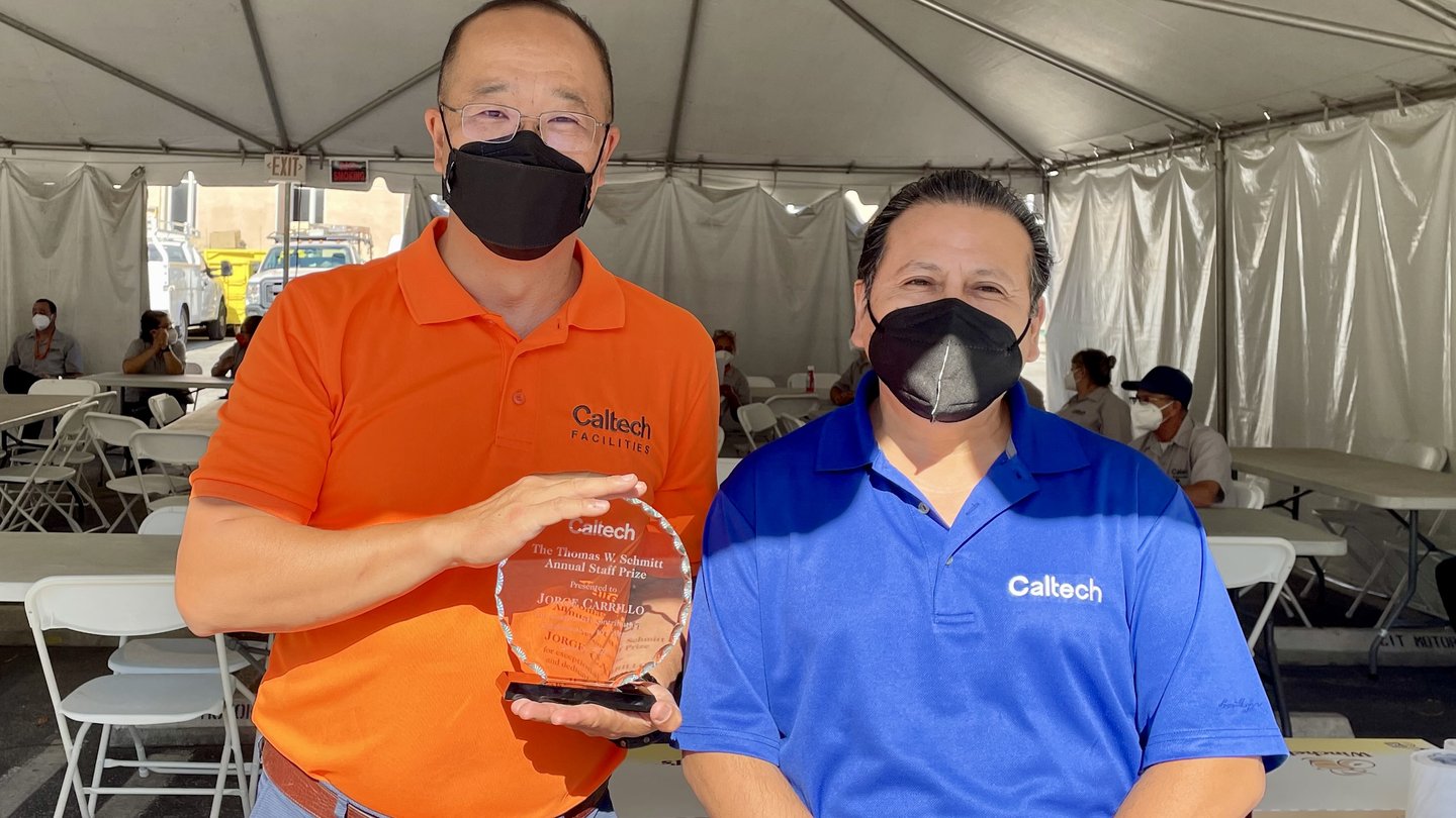 two men standing in a large white tent in shirts with collars, one shirt bright blue and one shirt bright orange. Both men are wearing black masks. One man is holding a round award with engraving