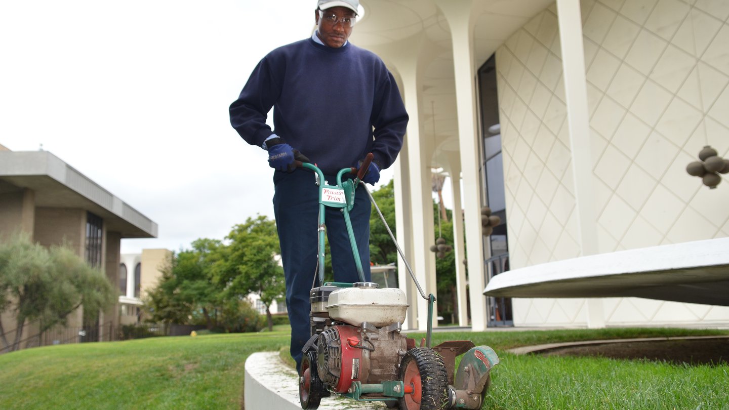 man in dark blue long-sleeved shirt and long dark-blue pants is pushing a machine to cut the edge of grass. Behind him is a tall, round, white building with white columns
