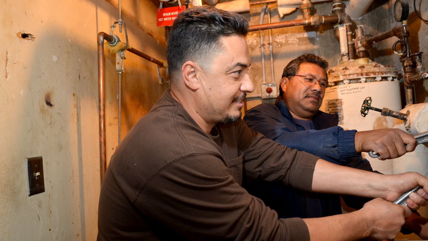 two men in a machine room with copper pipes. They are both holding wrenches. Man in foreground is wearing a dark brown shirt. Man in background has glasses and is wearing a dark blue shirt.