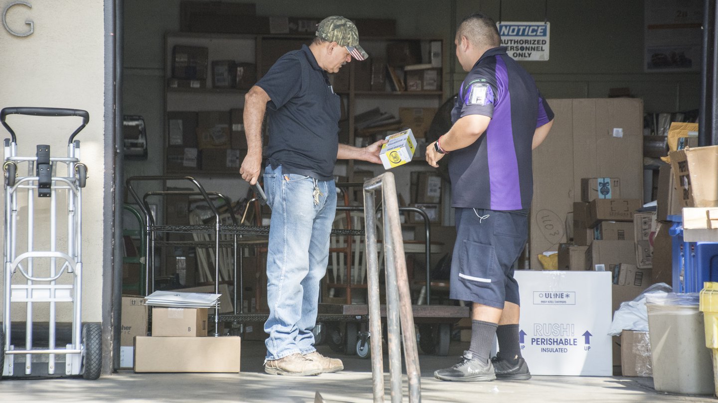 a man in jeans and a dark blue shirt, wearing a baseball cap, is holding a small box. Another man has dark blue shorts with a dark blue shirt with a purple vertical stripe. Behind them are many boxes and small wheeled carts for pushing things