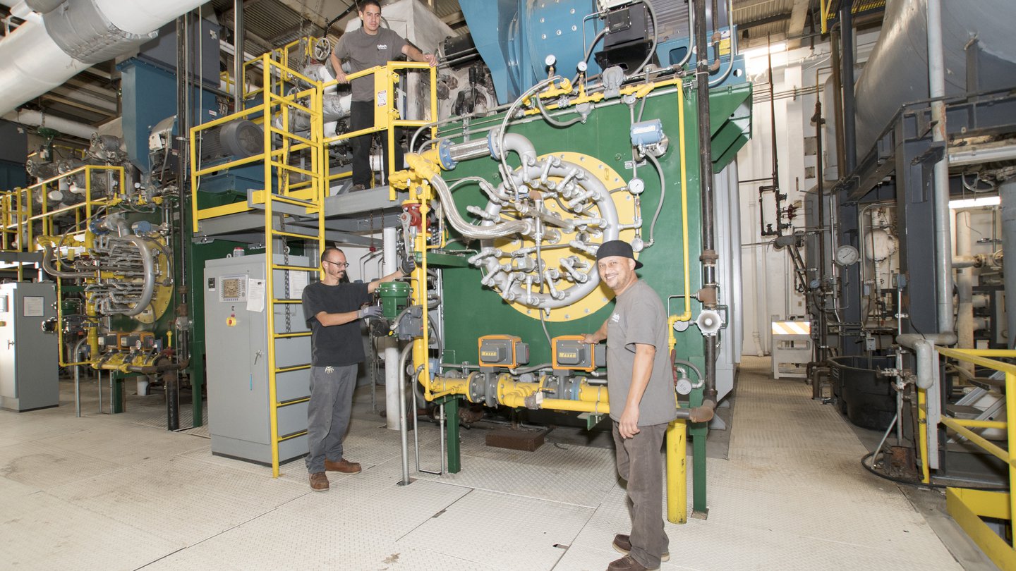 two men standing in front of a large green machine with yellow and silver pipes attached. One man has a black cap on backwards and a tan shirt; the other man has a black shirt. The machine is square and is twice as tall as the two men.