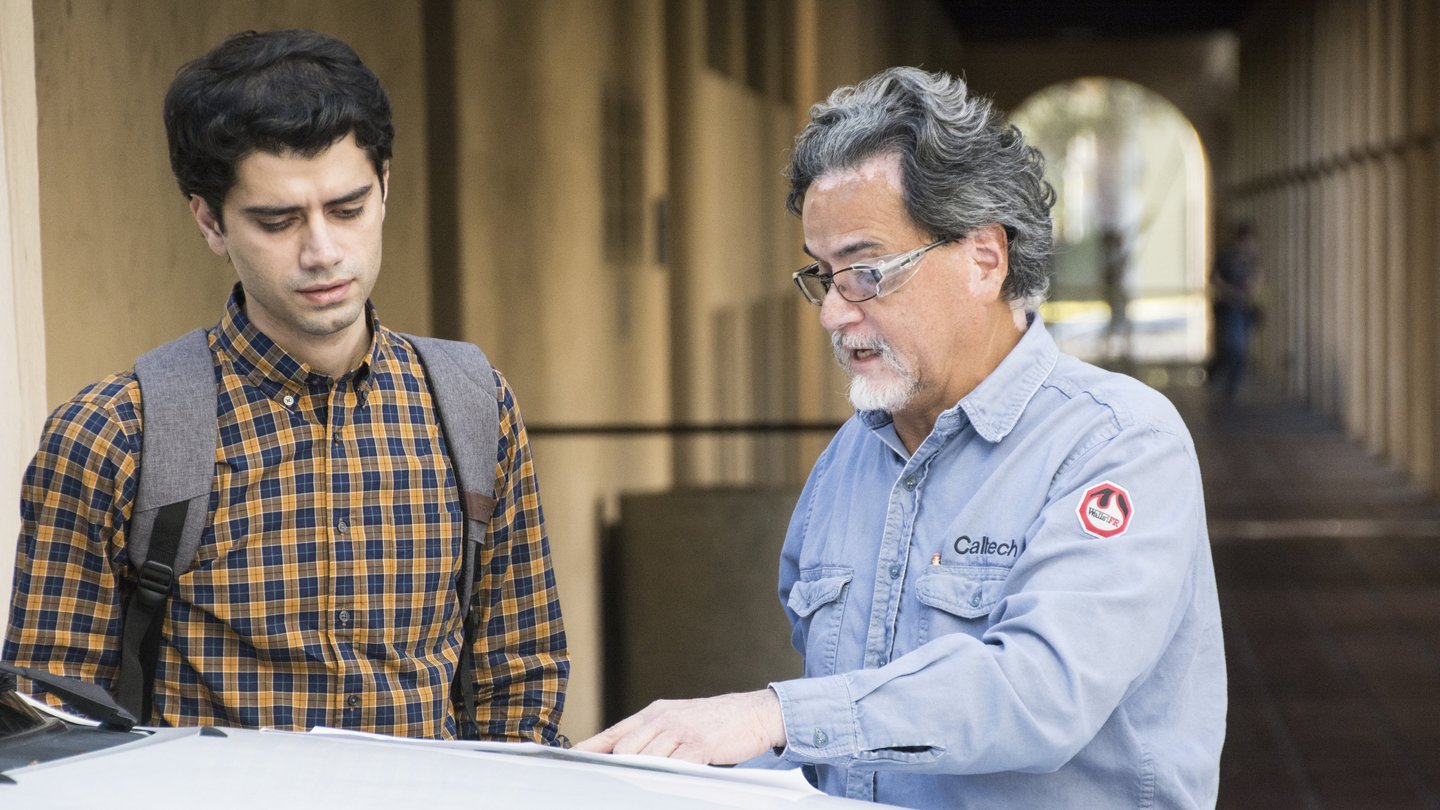Facilities worker in blue shirt pointing to document alongside Student in brown and black checked shirt