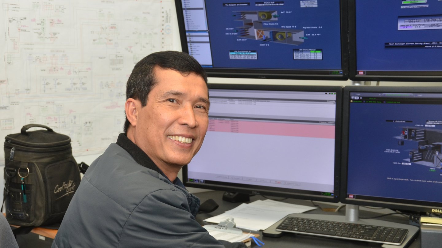 Paul Ayala from Service Center sitting in front of computer with graphical images on the screen. There is a white board in the background with figures written on it.