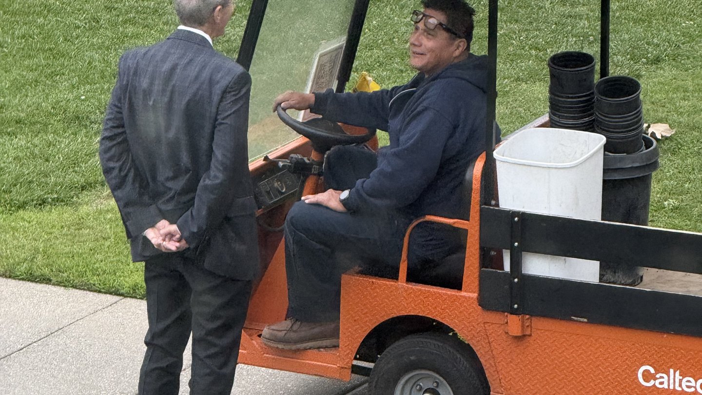 Man in gray suit with hands behind his back stands next to man driving orange utility cart