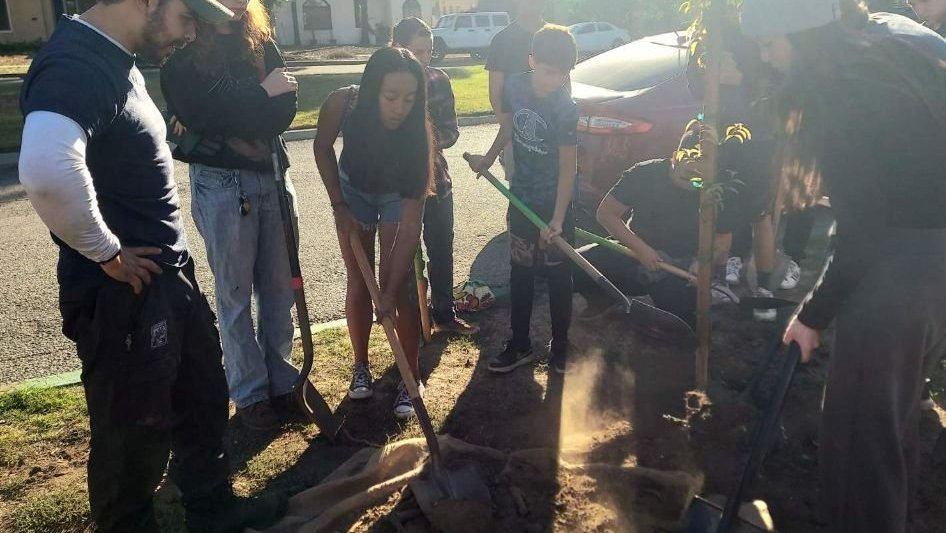 A group of young people is standing around a new tree, holding shovels. One is a young boy and one a young girl. The people are all wearing dark blue shirts.