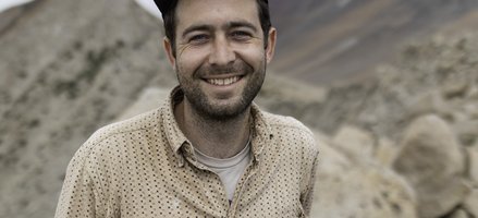A man with a beard wearing a baseball cap and a button down shirt smiling in front of a tan and red rocky background.