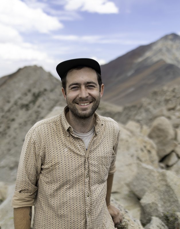 A man with a beard wearing a baseball cap and a button down shirt smiling in front of a tan and red rocky background.
