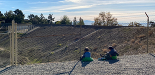 Two children sitting on the ground overlooking a debris catchment basin on a sunny day.