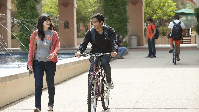 A student on a bicycle talking to another student.