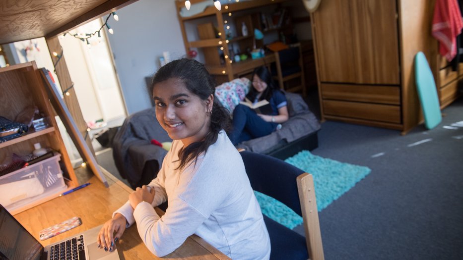 Student studying at her desk and roommate reading book in background.