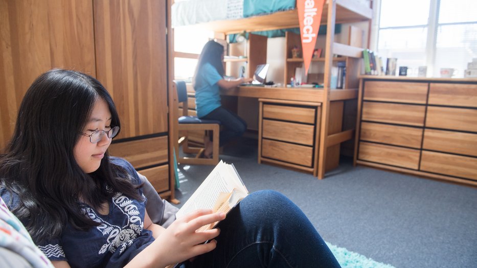 Student reading a book in Caltech's Avery House, roommate studying on laptop at her desk in the background.