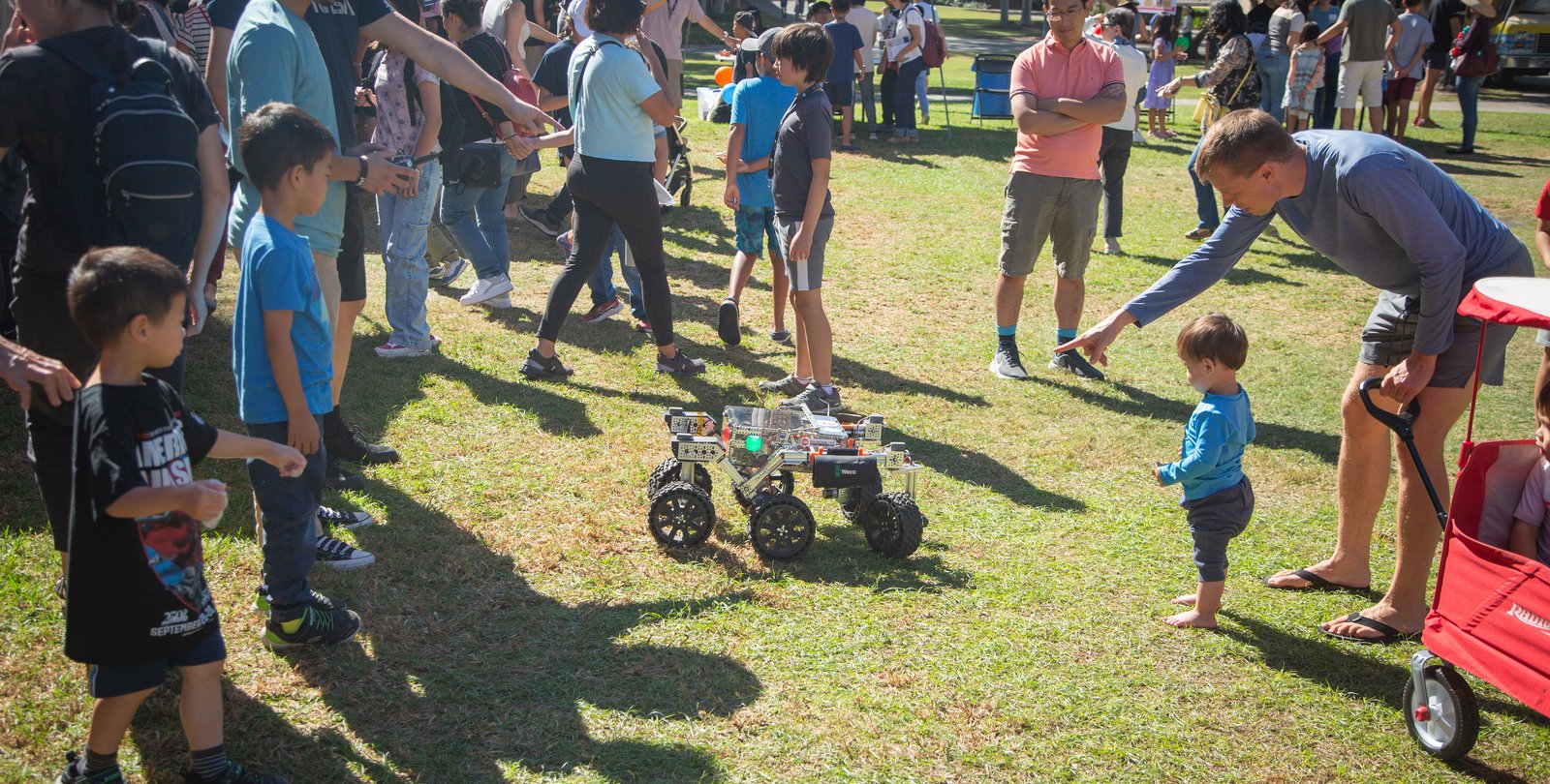 A group of parents and children observe a robotics team project demonstration